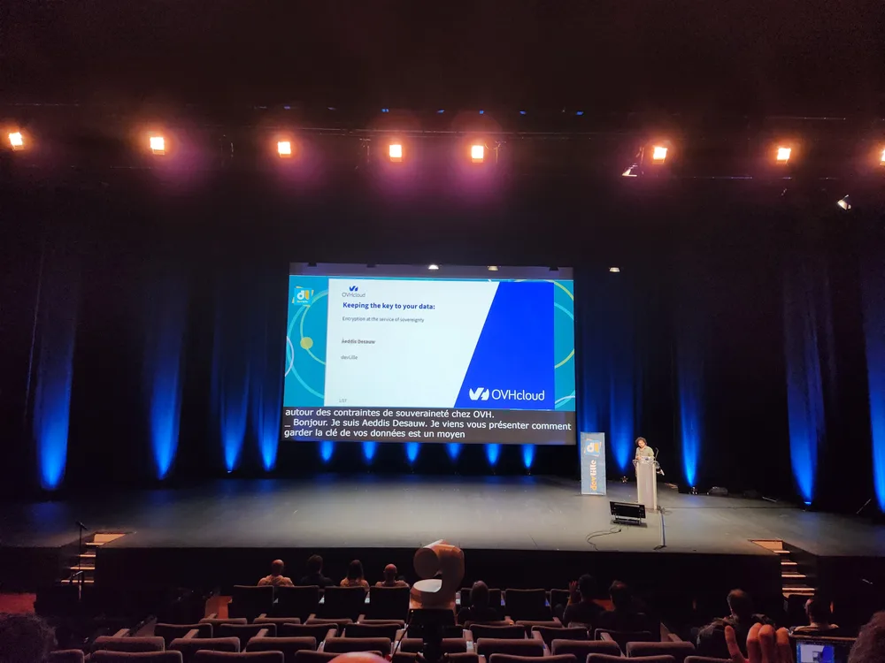 Une vue depuis le public de la conférence sur la scène du grand théatre au Grand Palais de Lille. Le présentateur se tient à un pupitre à droite, à côté d'un grand écran central affichant une présentation d'OVHcloud intitulée « Keeping the key to your data: Encryption at the service of sovereignty » par Aeddis Desauw. La scène est éclairée par des projecteurs bleus et chauds.