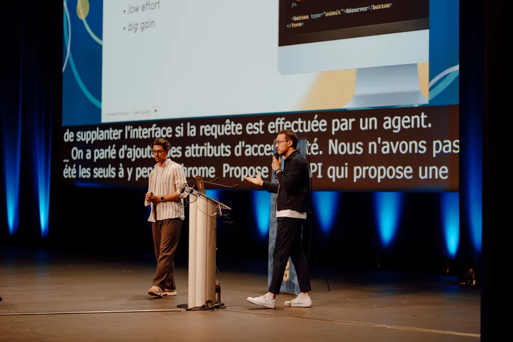 Jonathan & Julien sur la scène du Grand Théatre du grand palais dans le cadre du  DevLille 2026, l'un marchant près du pupitre, l'autre parlant au micro, devant un grand écran affichant du code HTML et des sous-titres en direct.