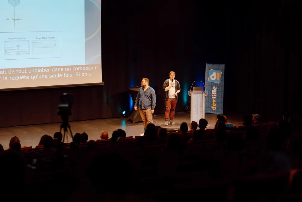 Lucas et Delphin debout sur la scène Marie Curie au Grand Palais dans le cadre du DevLille 2026 face au public, devant un slide montrant les maquettes « Page liste Recettes » et « Page détail Recette ».