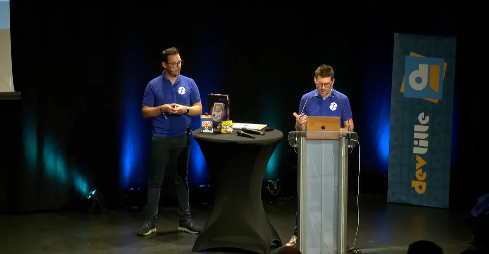 Quantin et Ludovic  en polo bleu Zenika présentent sur scène au DevLille, l'un au pupitre avec un ordinateur portable, l'autre à côté d'une table avec une console de jeux rétro.